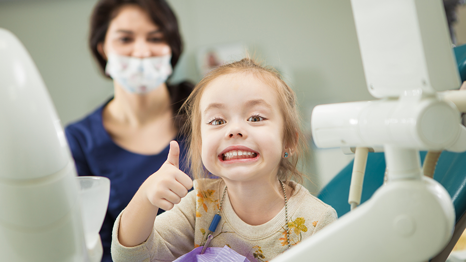 En strålandeglad flicka i tandläkarstolen visar tummen upp